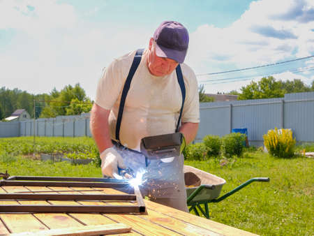 A senior man in a cap welds metal pipes on a table in the yard of a country house. The master works with electric arc welding.の写真素材