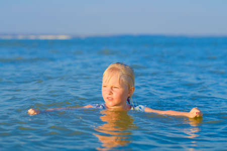 A blonde child girl bathes in the sea on a sunny summer day. The head of a happy child sticks out of the water.の写真素材
