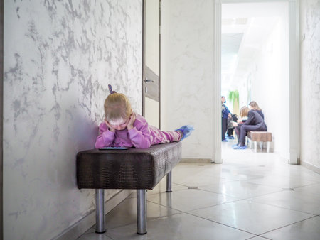 A little child girl lies on a couch in a clinic and watches a cartoon on a smartphone. Digital entertainment for kids.の写真素材