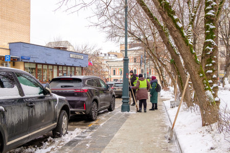 Moscow. Russia. February 25, 2023. Utility workers in yellow-green vests remove snow from the sidewalk on a city street on a cloudy winter day.のeditorial素材