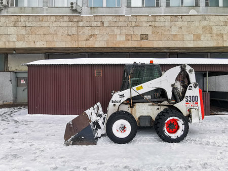 Moscow. Russia. March 6, 2023. A small snowplow Bobcat backhoe loader stands in the snow against the side of a building. Snowflakes fly in the air.のeditorial素材