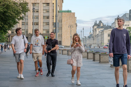 Moscow. Russia. July 1, 2023. A young beautiful girl in a light dress walks along a city street in summer. The girl looks at the smartphone screen on the go.のeditorial素材