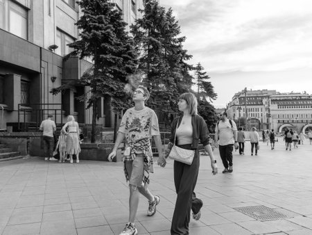 A young couple is walking along a city street and smoking a vape. Black and white photo. Moscow. Russia. July 1, 2023.のeditorial素材