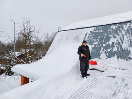 An elderly man with a snow shovel on the roof of a country house on a winter day. Clearing the roof of snow during snowfall.の写真素材