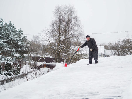 An elderly gray-haired man moves snow from the roof with a red shovel on a winter day. Clearing snow from the roof of a country house.の写真素材