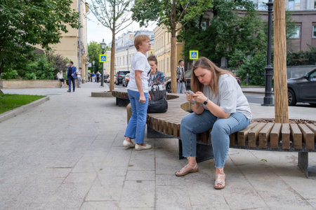 A plump young woman with a smartphone in her hands sits on a bench in the city on a summer day. Moscow. Russia. June 30, 2024.のeditorial素材