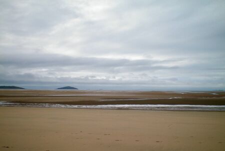 A receding tide has left a large amount of sand exposed on a beach. Some water remains. Two small hills are visible in the distance, and two tankers are just visible on the horizon. The sky is grey and overcast.の写真素材