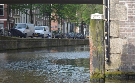 A wooden log marking the corner of a canal in Amsterdam. The wood is chipped and warped with age. It is beside a brick-wall, and the bottom of a bridge frames the view. The image has a shallow depth of field, with the side of the canal and a row of cars, の写真素材