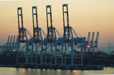 An industrial site for loading cargo ships. Cargo containers fill the dock. The colours of dawn fill the sky, and are reflected in the water.の写真素材
