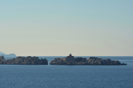 The red roof and light of a light house rise above an island of rocks. The rocks are surrounded by ocean. The sky is clear.の写真素材