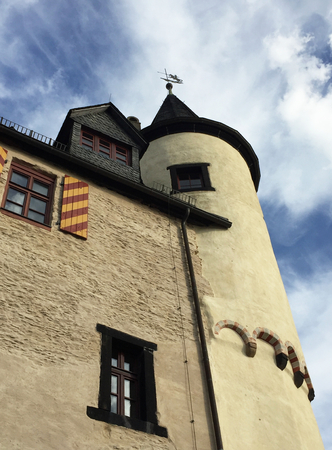 A tower with a turret is seen rising into a blue sky with white clouds. A weather vane is on the roof. Windows are in the wall of the castle, one with red and yellow striped shutters.のeditorial素材