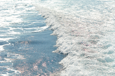 The wash from a boat bubbles white over the blue water. It is tinged red from floating algae.の写真素材