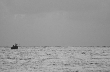 A small fishing vessel is silhouetted against the horizon. The sky is overcast, and the sea a silver grey. Two people can just be seen. The photograph is in black and white.の写真素材
