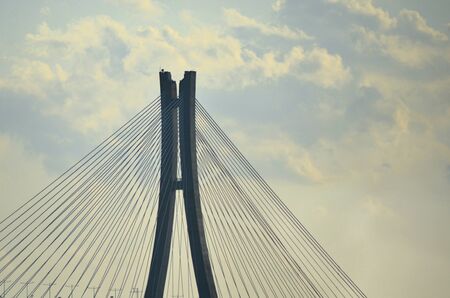 Two parts of a pylon rise vertically, with supportive metal strusts radiating off them in a diagonal pattern. The sky is pale blue, with white and grey clouds.の写真素材