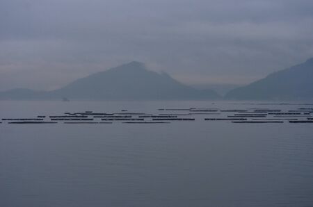 A bay at sunset, filled with mist. Mountains are on the far shore. Wooden floats used for fishing and oyster farming are in the foreground.の写真素材