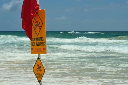 A yellow sign with a red flag is warning against swimming on a beach. Rough surf is the background...