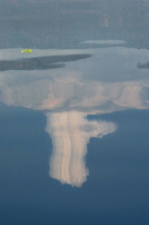 An unusually tall white cloud is reflected in the blue waters of a lake. Other clouds and a mountain...