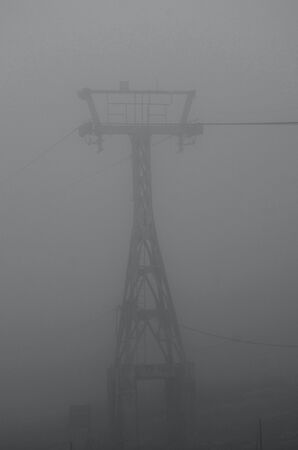 The tower for a cable car is just visible through a mist which fills a valley. The wires disappear into the gloom. The photogrpah is in black and white.の写真素材