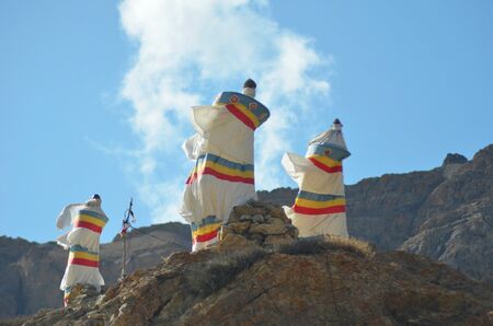 Three shrines made of white fabric rest on top of a rocky outcrop of barren hills in Ladakh. They have red, yellow and blue stripes, and are bending in the wind. The sky is blue with white clouds.の写真素材