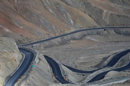 The black colour of a winding road contrast with the red and browns of the surrounding mountain slopes in the Western Himalayas. A few workmen are visible as tiny figures, dwarfed by the size of the mountains.の写真素材