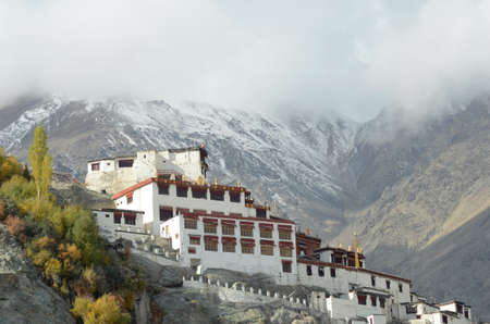 The white buildings of Diskit Monastery are built into the slope of the surrounding mountains. Snow and mist cover the top of the mountains, and some trees are beside the monastery.のeditorial素材
