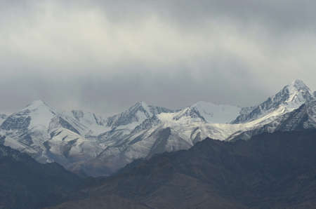 Snow covered mountain peaks rise into grey storm clouds. A shaft of sunlight falls onto a patch of snow. Rocky mountain slopes are in the foreground.の写真素材
