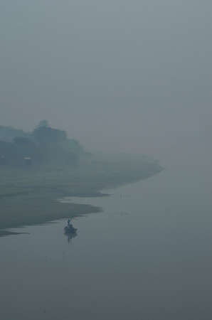 A fisherman standing in his boat, using a pole to steer it. He can barely be seen against the vastness of the lake, which is covered in mist. Trees on the shore are to one side, and cables crossing the water are just visible.の写真素材