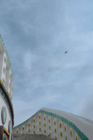A helicopter is seen from below, against a blue sky with white clouds. Beneath it are the steps of a Buddhist temple, painted white with gold decorations.の写真素材