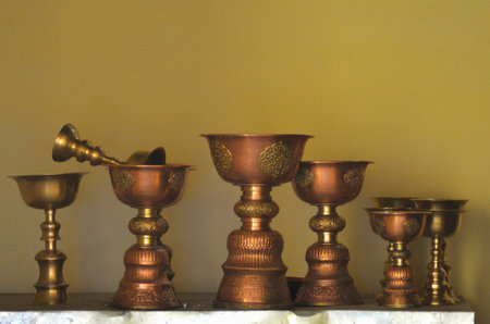 Golden cups used in Buddhist religious ceremonies standing on a shelf in a temple. Some are plain, while others carved details. The wall behind is dark yellow, giving the photo golden tones.の写真素材