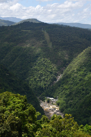 A clearing is visible in a steep valley of a rain forest. Wires stretch from the building to electricity poles running up and across the hills. The sky is blue with grey and white clouds.の写真素材