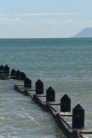The rising tide has flooded an ocean pool. Only the top of the wooden posts, with netting attached, are visible. The water stretches to the horizon. Hills are in the distance, and the sky is blue with white clouds.の写真素材