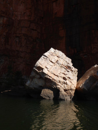 A large boulder lies in a river. Sunlight reflects off  the surface, leaving the rest of the photograph dark in contrast. The cliff behind is faintly visible.の写真素材