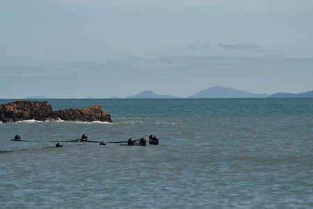 The poles of a netted ocean bath are just visible in a high tide. Rocks are behind the pool, and hills can be seen across the bay. The sky is blue, with faint clouds.の写真素材