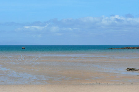 A sandy beach stretches into a blue sea. There is a small boat with a flat top, and a rock shelf. Pale blue clouds are on the horizon.の写真素材
