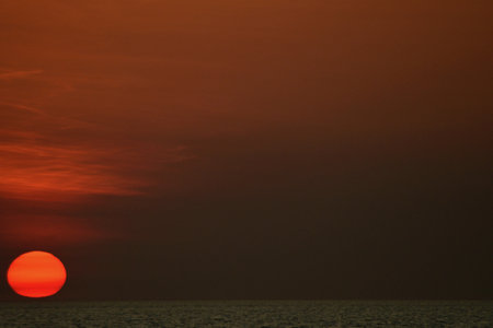 The sun is setting into a dark sea. The clouds above it are a dark orange, while the rest of the sky is dark. The sun is a vibrant orange with bands of red.の写真素材