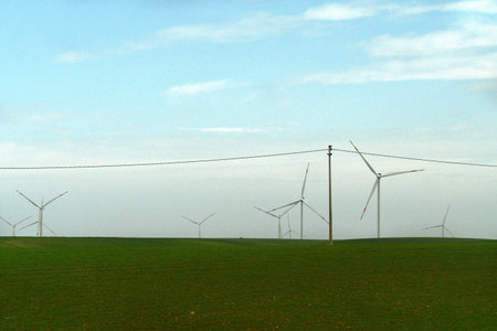 White wind turbines are on a green field, which also has a pole with an electricity wire. The sky is blue, with white clouds.の写真素材