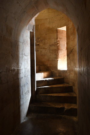 Worn stone stairs form a spiral staircase in the turret of a castle. Sunlight falls through a...