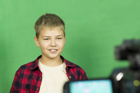 Young boy blogger records video on a green background. A boy blogger records a video on a green background at home on a video camera. A blogger is sitting on a chair with a laptopの写真素材
