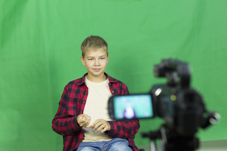 Young boy blogger records video on a green background. A boy blogger records a video on a green background at home on a video camera. A blogger is sitting on a chair with a laptopの写真素材