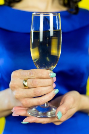 A girl with a glass of champagne smiles in front of the camera. Beautiful brunette girl in a blue dress drinking champagne on a yellow backgroundの写真素材