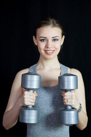 The athlete is engaged with heavy dumbbells on a black background. A beautiful girl in gray portable form raises heavy gray dumbbells.の写真素材