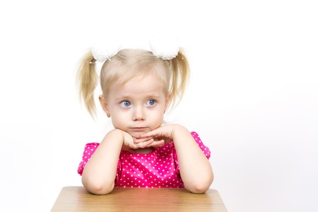 A beautiful young blond girl leans her elbows on a chair and puts her head on her hands. Little girl 3 years old on a white background put her head on her hands.の写真素材