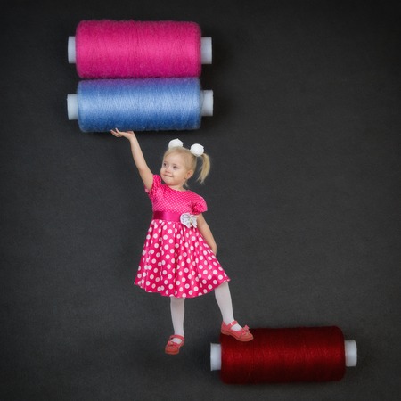 A small child in the world of big people. A beautiful girl holds two huge coils with sewing threads...