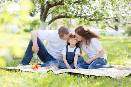 A happy family of three people at a picnic. Parents kissing their beautiful daughter sitting under a tree.の写真素材