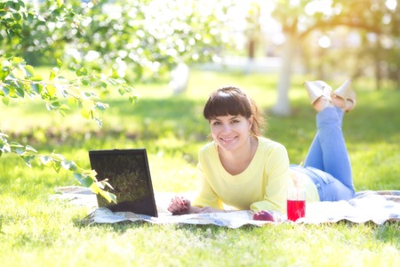 Girl freelancer works in the open air. The brunette lies on the grass in the park. The herb is her breakfast - juice, fruit, biscuits.の写真素材