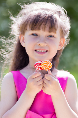 A girl in a red dress with sweets in her hands. Beautiful little girl is playing with a big sweet candy in the city park.の写真素材