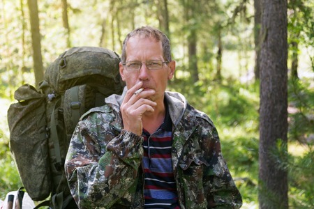 A man with a backpack smokes cigarettes. The tourist on vacation after a hike in the forest.の写真素材