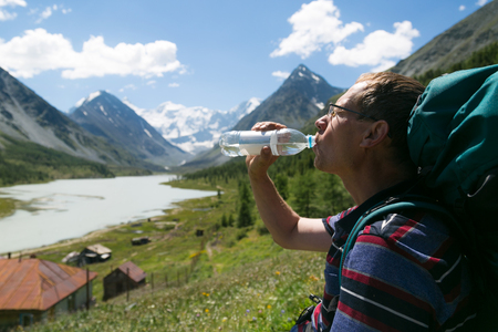 A man drinks water from a bottle in the background of the mountains. Tired tourist quenches thirst from the bottle, clean water, ecology, beauty of nature.の写真素材