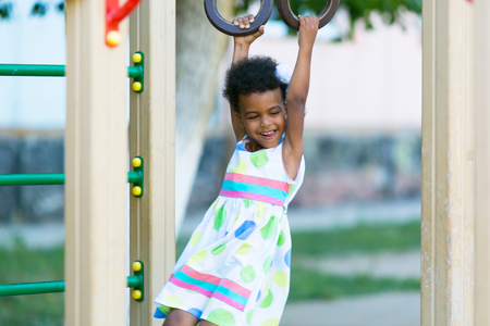 The girl is hanging on the sports rings. African American girl plays on a sports playground.の写真素材