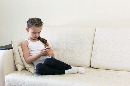 A girl with a mobile phone on the couch. A child plays with the phone while sitting in a room on the couch.の写真素材
